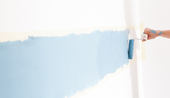 Kuvassa näkyy maalitahrainen käsi vetämässä vaaleansinistä maaliraitaa valkoiseen seinään telalla.