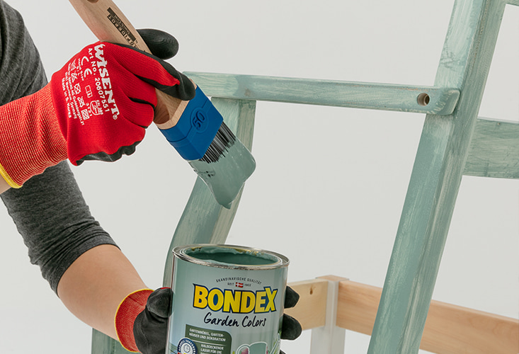 Kuvassa näkyy punaisilla työhanskoilla verhoillut kädet, joissa toisessa on salvian vihreään maaliin kastettu maalipensseli ja toisessa maalipurkki. Taustalla näkyy osittain maalattu tuoli, jonka pohjaväri on vaalea puu.